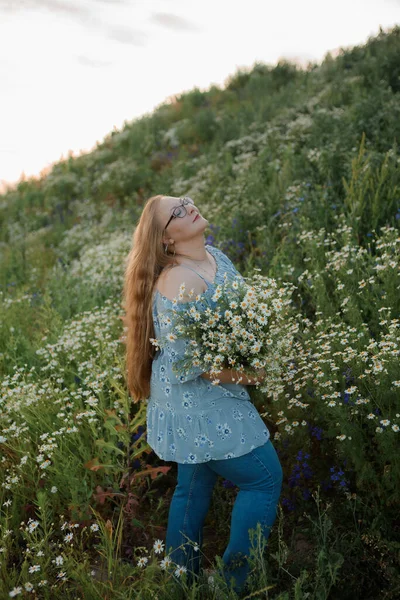 Fotoğrafta uzun sarı saçlı bir kız tarlada yürüyor ve küçük beyaz bir papatya buketi topluyor.