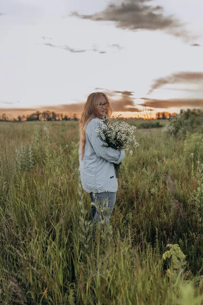 Bir kız tarlada yürüyor, ve bir buket küçük beyaz papatya çiçeğiyle poz veriyor, arka planda yeşil bir tarla ve turuncu bir gün batımı, uzun sarı saçlı bir kız