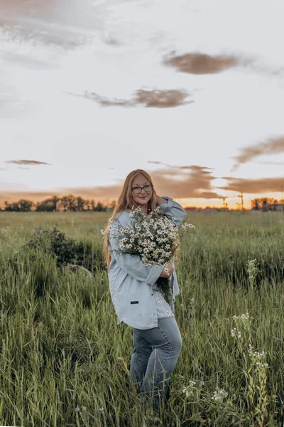 Bir kız tarlada yürüyor, ve bir buket küçük beyaz papatya çiçeğiyle poz veriyor, arka planda yeşil bir tarla ve turuncu bir gün batımı, uzun sarı saçlı bir kız