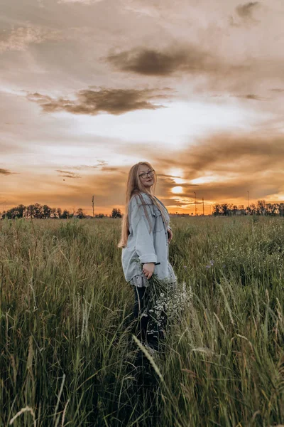 Bir kız tarlada yürüyor, ve bir buket küçük beyaz papatya çiçeğiyle poz veriyor, arka planda yeşil bir tarla ve turuncu bir gün batımı, uzun sarı saçlı bir kız