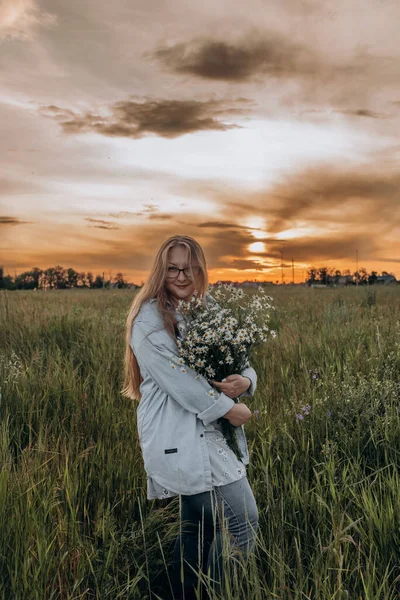 Bir kız tarlada yürüyor, ve bir buket küçük beyaz papatya çiçeğiyle poz veriyor, arka planda yeşil bir tarla ve turuncu bir gün batımı, uzun sarı saçlı bir kız