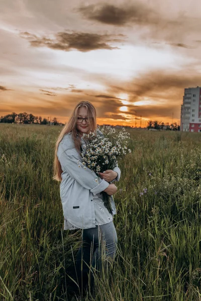 Bir kız tarlada yürüyor, ve bir buket küçük beyaz papatya çiçeğiyle poz veriyor, arka planda yeşil bir tarla ve turuncu bir gün batımı, uzun sarı saçlı bir kız