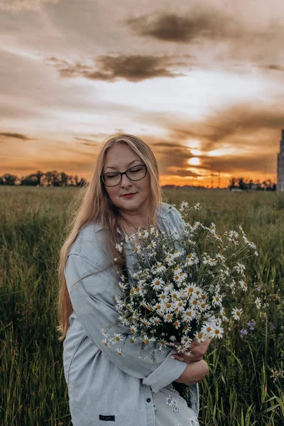 Bir kız tarlada yürüyor, ve bir buket küçük beyaz papatya çiçeğiyle poz veriyor, arka planda yeşil bir tarla ve turuncu bir gün batımı, uzun sarı saçlı bir kız
