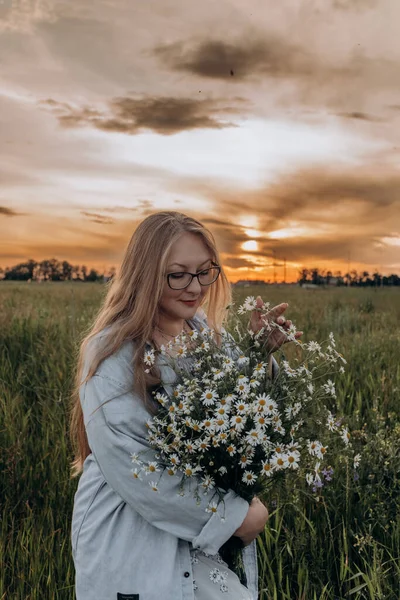 Bir kız tarlada yürüyor, ve bir buket küçük beyaz papatya çiçeğiyle poz veriyor, arka planda yeşil bir tarla ve turuncu bir gün batımı, uzun sarı saçlı bir kız