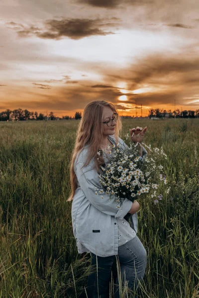 Bir kız tarlada yürüyor, ve bir buket küçük beyaz papatya çiçeğiyle poz veriyor, arka planda yeşil bir tarla ve turuncu bir gün batımı, uzun sarı saçlı bir kız