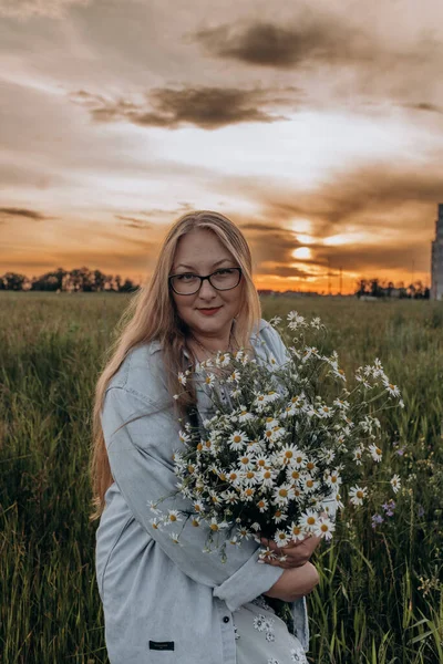 Bir kız tarlada yürüyor, ve bir buket küçük beyaz papatya çiçeğiyle poz veriyor, arka planda yeşil bir tarla ve turuncu bir gün batımı, uzun sarı saçlı bir kız