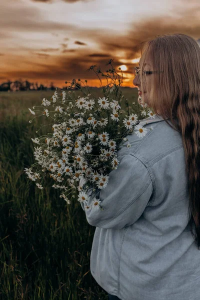 Bir kız tarlada yürüyor, ve bir buket küçük beyaz papatya çiçeğiyle poz veriyor, arka planda yeşil bir tarla ve turuncu bir gün batımı, uzun sarı saçlı bir kız