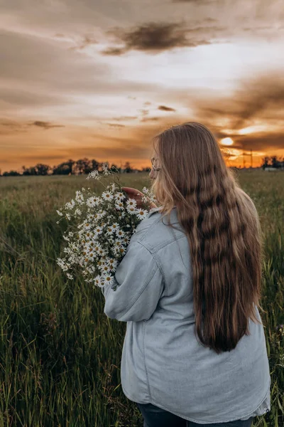Bir kız tarlada yürüyor, ve bir buket küçük beyaz papatya çiçeğiyle poz veriyor, arka planda yeşil bir tarla ve turuncu bir gün batımı, uzun sarı saçlı bir kız