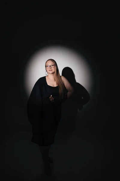 Siyah elbiseli, siyah ceketli, uzun sarı saçlı, siyah arkaplanda fotoğraf çeken bir kızın moda fotoğrafı.