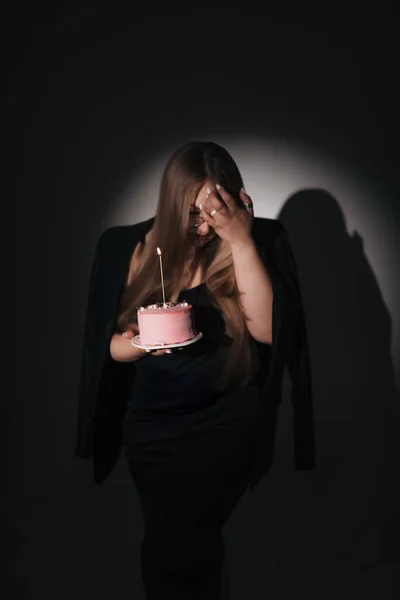 Siyah elbiseli, siyah ceketli, uzun sarı saçlı, fotoğraf stüdyosundaki siyah arka planda fotoğraf çeken, doğum günü pastası olan bir kızın moda fotoğrafı.