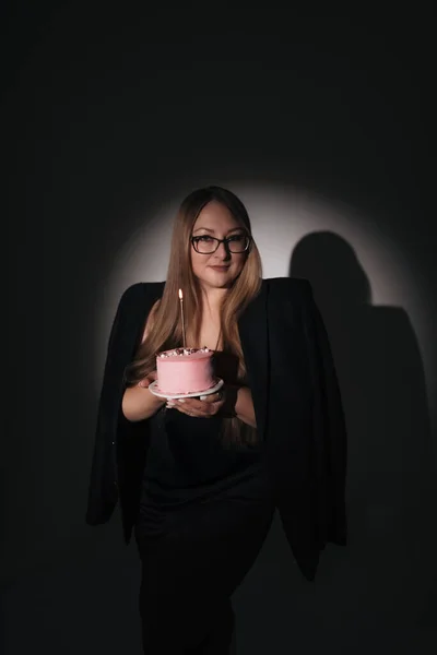 Siyah elbiseli, siyah ceketli, uzun sarı saçlı, fotoğraf stüdyosundaki siyah arka planda fotoğraf çeken, doğum günü pastası olan bir kızın moda fotoğrafı.