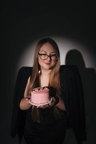 Siyah elbiseli, siyah ceketli, uzun sarı saçlı, fotoğraf stüdyosundaki siyah arka planda fotoğraf çeken, doğum günü pastası olan bir kızın moda fotoğrafı.