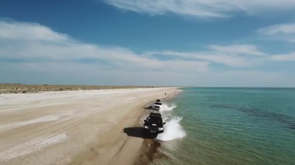 Los coches conducen a lo largo de la playa de arena creando aerosoles de agua. Vista aérea — Vídeos de Stock