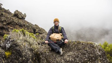 Renkli şapka içinde dağ turistik bir taş üzerinde oturan ve gülümseyerek. El çantası ve sopa üzerinde