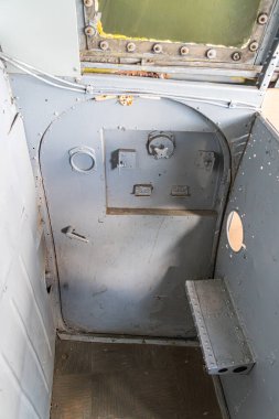 Kıvrımlı duvarları olan küçük bir kamara, muhtemelen küçük bir tekne ya da uçak Benç benzeri bir yapı, kapı, panel, kontrol tahtası Dünya tonları, sessiz mavi, loş ışıklandırma, ince gölgeler Cand