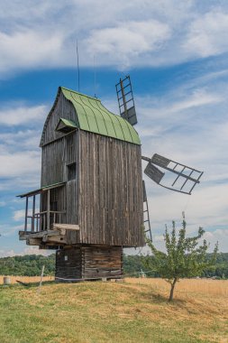 Çimenli bir yamaçta yer alan eski bir Orta Avrupa köyünde, sazdan bir çatı ve aşağıdaki yemyeşil çimlerin üzerinde küçük ahşap bir yapı, uzaktaki dağınık ağaçlar, Overca