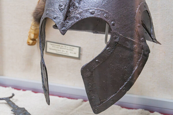 Close-up image of an Iron Age helmet, a weathered artifact with a brow guard and eye slots Displayed against a light-colored museum backdrop, the object is highlighted by soft shadows and even illum
