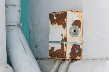 Beton bir duvardaki paslı metal panel, aşınmış ve soyulmuş boya izleri, sağ üst köşedeki virajda sürgülenmiş dikey metal boru, kapalı ya da kapalı olduğunu ima eden endüstriyel bir sahne olduğunu gösteriyor.