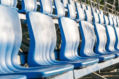 Metal tribünlerdeki mavi stadyum koltukları, sıra sıra dizilmiş koltuklar, boş yer yok, mavi ve metalik gri renkler, hatta sert gölgeler olmadan aydınlatma, beklenti