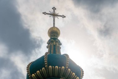 Mavi kubbeli bir kilise kulesinin tepesinde, bulutlu gökyüzüne karşı duran altın bir haç.