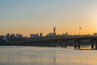 Köprü ve kentsel binalarla Kyiv nehrinin üzerinde ılık gün batımı, durgun su gökyüzünü yansıtıyor