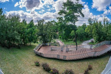 Alacakaranlık sırasında modern bir şehir parkı sahnesi, çok katlı ahşap bir pavyon ve yeşil ağaçların arasında dolambaçlı bir yol. Yumuşak, yayılmış ışık mavi-sarı eğimli gökyüzü.