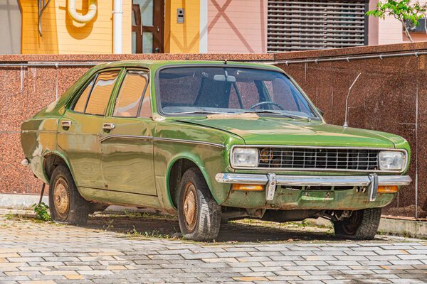 Vintage green car, mid-20th century design, angled on cobblestone street, urban residential background, faded bodywork, chipped and rusted paint, round headlights, visible engine compartment Natural
