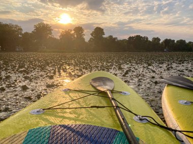 Turuncu ve sarı bir gökyüzüne karşı, nilüferlerle kaplı bir su üzerinde iki canlı kanoyla sakin bir günbatımı sahnesi.