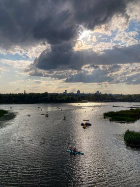 Kyiv 'de bir köprü, ağaçlar, küçük tekneler sarı kano, kırmızı kano, bulutlu gökyüzü, ara sıra güneş ışığı, mavi ve yeşil su, doğal renkler, gündüz Kyiv, Ukrayna 08-17-2025