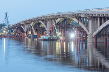 Kyiv, Ukrayna 'da kısmen sökülmüş beton bir köprünün gece inşaatı iskele ve inşaat ekipmanları, büyük sarı vinçler, sakin su Kyiv, Ukrayna 06-01-2025