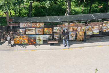 Bir adam, Kyivs Sanatçılar Sokağı 'ndaki renkli resimlerin açık havada sergilendiği bir gösterinin ortasında gündüz vakti bir tepe örtüsünün altında çeşitli konuları ve stilleri gösteriyor.