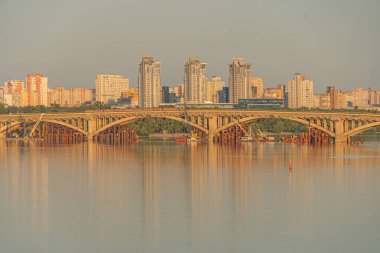 Gün batımında Kyiv şehri manzarası, ılık ışığı yansıtan sakin su, nehir üzerindeki kemerli köprü, yemyeşil ağaçlar, katmanlı şehir silueti, toprak rengi, yumuşak ışıklandırma, Kyiv, Ukrayna 07-05-2025