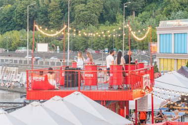 Alacakaranlıkta limanda fener ve sicim ışıkları olan canlı kırmızı gıda standı, yakınlardaki insanlar yiyor veya sosyalleşiyor, ek şemsiyeler, arka planda kayıklar, sıcak lig Kyiv, Ukrayna 07-06-2025