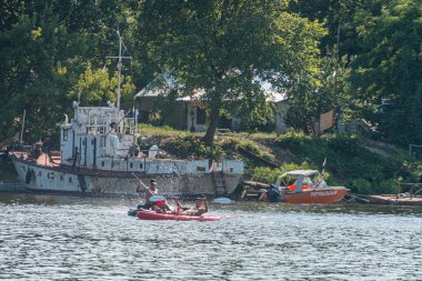 Kırmızı kanolu biri sağa doğru kürek çekiyor, turuncu sürat teknesi ahşap iskeleye yanaşmış, sürat teknesinde ikinci kişi, yemyeşil arka plan, ılık ışık, öğleden sonra Kyiv, Ukrayna 07-12-2025