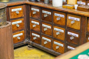 Antika eczane dolabı, birden fazla çekmeceli, beyaz etiketli organize bir masa, açık kahverengi ahşap kaplama, sağlam bacaklar, metal lamba, süslü taban, kıvrımlı gölge, sıcak parıltı, yeşil çömlek kavanozu, üniversite.