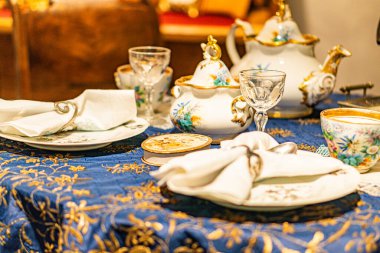 Kaliteli Çin porselenleri, kristaller, gösterişli şal desenli masa örtüsü, karmaşık çiçek tabakları, hassas bardaklar, çatal bıçak takımı, çaydanlık, yumuşak ışıklandırma, rafine ve ele