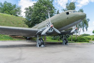Beyaz işaretli klasik zeytin yeşili askeri uçak, belirgin özellikli pervaneler, kokpit kapısı açık, parçalı bulutlu gökyüzü, manikürlü çimenler ve ağaçlar, potansiyel müze veya sergi alanı