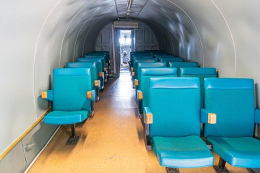 Bir uçak kabini ya da denizaltıyı andıran tünel benzeri bir yapının içinde, her iki tarafında da mavi koltuklar, kabza, yapay ışıklandırma gölgeleri, görünür insanlar yok.