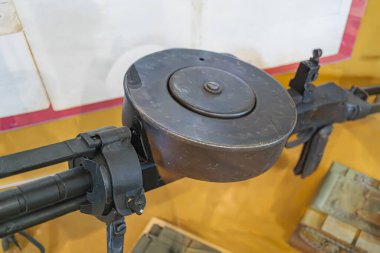 Diğer askeri eserlerle çevrili bir müze ortamında sergilenen antika bir tüfek koyu renkte ve karşıt sarı bir masaya yerleştirilmiş.