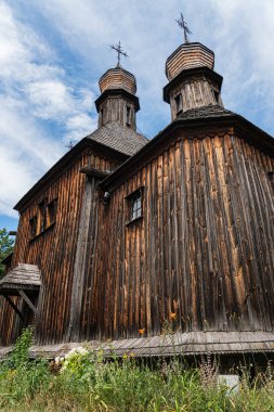 Orta Avrupa kırsal kesimindeki eski Ortodoks ahşap kilise, parçalı bulutlu gökyüzünün altında, birden fazla çan kulesi ve haçı olan karanlık ahşap cephe, altında aşırı büyümüş çimenler.