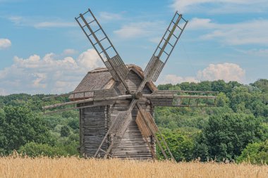 Açık alanda büyük ahşap yel değirmeni, sazdan çatı, çoklu yelkenler, parçalı bulutlu gökyüzü, yemyeşil