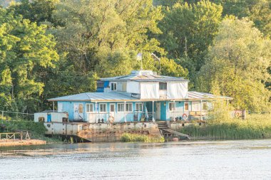 Sudaki canlı mavi yüzen ev, muhtemelen bir nehir ya da göl yemyeşil ve rıhtım. Yandaki iki pencere ve bir yan kapıdan ulaşılabilir.