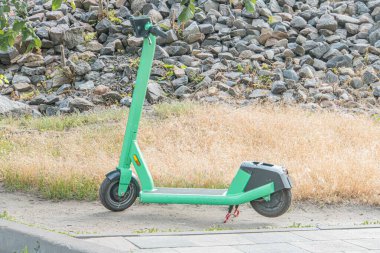Açık hava kentsel sahnesinde park etmiş elektrikli scooter siyah tekerlekler, koyu mavi koltuk, kumlu ya da kayalık zeminde açık yeşil vücut