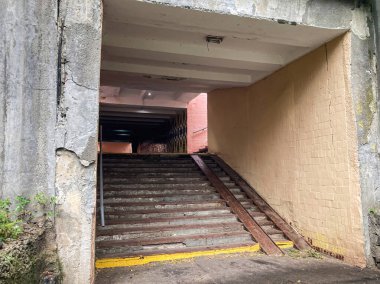Kapalı girişi olan terk edilmiş alt geçit, aşınmış merdivenler ve sarı çizgili beton duvarlar metal kirişlerle desteklenen çatlaklar gösteriyor.
