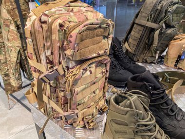 Perakende bir ortamda, büyük bir kamuflaj sırt çantası ve uyumlu botların etrafında toplanmış askeri eşyaların sergilenmesi.