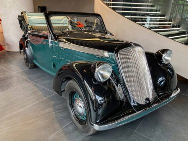 Beyaz aksanlı, krom detaylı, kapalı sergi alanı veya galeride sergilenen 1930 model koyu mavi bir araba. Araç 20. yüzyılın ortalarında yuvarlak bir vücut stiline sahip.