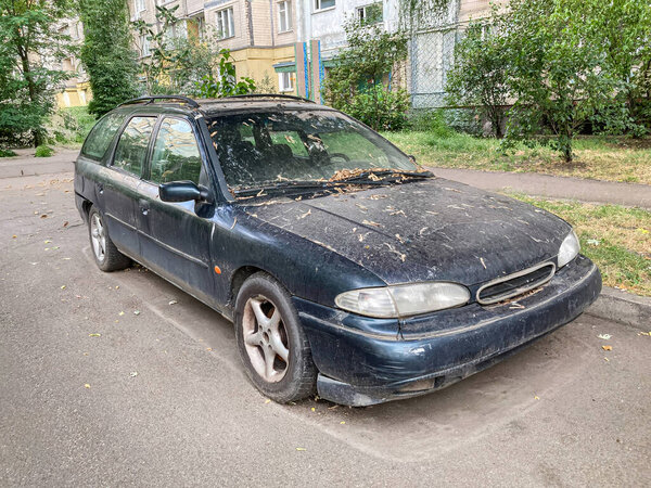 An older blue car with accumulated leaves on the hood is parked on a quiet, muted residential street The car and buildings suggest an urban neighborhood Soft natural lighting creates gentle shadows