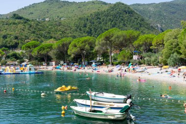 08.23.2025 - Medveja, Croatia: Sunny beach with swimmers, boats, and inflatables by green hills and pine trees .