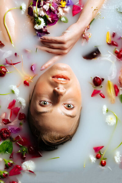 A young woman with a stunning hairstyle is in a bath filled with milk and flowers. Woman in a milk bath with chamomiles, peonies, roses, and petals.