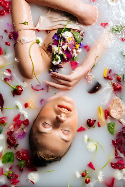 A young woman with a stunning hairstyle is in a bath filled with milk and flowers. Woman in a milk bath with chamomiles, peonies, roses, and petals.